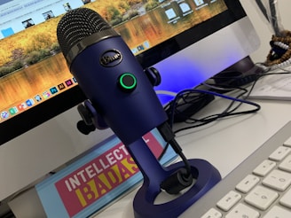 A blue microphone on a stand is placed in front of a computer monitor displaying various applications and a scenic wallpaper. The setup includes a keyboard and tangled cables. There is also a book or sign with the title 'INTELLECTUAL BADASS'.