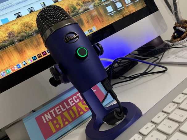 A blue microphone on a stand is placed in front of a computer monitor displaying various applications and a scenic wallpaper. The setup includes a keyboard and tangled cables. There is also a book or sign with the title 'INTELLECTUAL BADASS'.