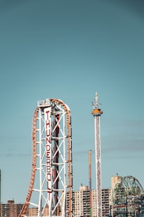 A roller coaster named Thunderbolt stands prominently with its loops and turns, alongside a tall thrill ride set against a backdrop of urban buildings and a Ferris wheel. The sky is clear, contributing to a sense of excitement and adventure.