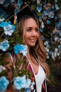 A woman wearing a graduation cap and gown is surrounded by vibrant blue flowers. Her hair is long and wavy, and she appears to be smiling with a joyful expression.