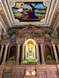 The image features an intricately decorated interior of a religious structure with ornate carvings on columns and walls. The ceiling showcases a vivid religious painting depicting an angel and a saint or prophet in a scene. Additionally, there is a prominent statue set in a niche surrounded by vegetation and candles.