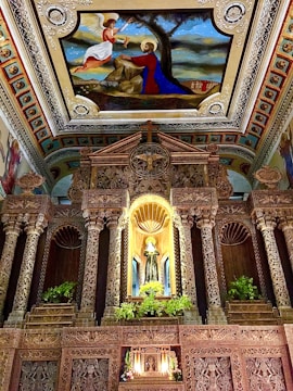 The image features an intricately decorated interior of a religious structure with ornate carvings on columns and walls. The ceiling showcases a vivid religious painting depicting an angel and a saint or prophet in a scene. Additionally, there is a prominent statue set in a niche surrounded by vegetation and candles.