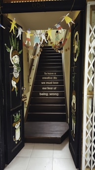 A staircase leading upwards is decorated with colorful paper cut-outs of people and various decorations hanging from the ceiling. The walls are dark with artistic illustrations of people and animals. A motivational quote is painted on the steps, and a small open sign hangs on the door frame.