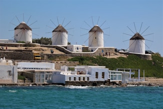 A coastal scene with several traditional white windmills featuring conical thatched roofs. The windmills are situated on a slightly elevated area covered in greenery. Below, near the shoreline, are white buildings with clear glass windows. The sea is visible in the foreground, with its waters a mix of blue and turquoise hues.