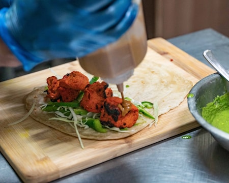 A gloved hand holds a squeeze bottle, applying sauce to a wrap filled with red grilled meat, shredded cabbage, and sliced green bell peppers on a wooden cutting board. A silver bowl containing a green sauce is visible on the side.