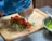 A gloved hand holds a squeeze bottle, applying sauce to a wrap filled with red grilled meat, shredded cabbage, and sliced green bell peppers on a wooden cutting board. A silver bowl containing a green sauce is visible on the side.