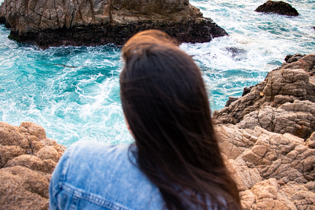 woman in blue denim jacket sitting on rock near body of water during daytime