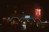 A dimly lit building displays neon signs, with 'Surfside' prominently illuminated in blue on the facade. The structure features ornate arches adorned with string lights and a glowing sign extends from the upper level. The overall atmosphere is nighttime, with people visible near the entrance and warm light spilling from the windows.