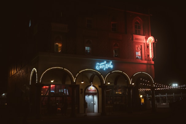 A dimly lit building displays neon signs, with 'Surfside' prominently illuminated in blue on the facade. The structure features ornate arches adorned with string lights and a glowing sign extends from the upper level. The overall atmosphere is nighttime, with people visible near the entrance and warm light spilling from the windows.