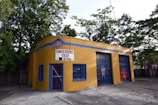 A small, yellow-brick building with blue accents is positioned near a cluster of trees. The front has a sign reading 'Joe's Emissions Test Waiting.' The building features a door with blue bars and two garage entrances. The ground is paved, and there's a fence visible in the background.