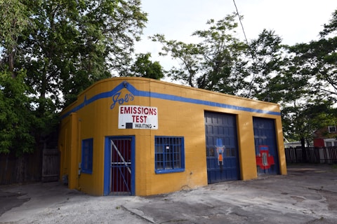 A small, yellow-brick building with blue accents is positioned near a cluster of trees. The front has a sign reading 'Joe's Emissions Test Waiting.' The building features a door with blue bars and two garage entrances. The ground is paved, and there's a fence visible in the background.