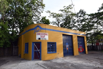 A small, yellow-brick building with blue accents is positioned near a cluster of trees. The front has a sign reading 'Joe's Emissions Test Waiting.' The building features a door with blue bars and two garage entrances. The ground is paved, and there's a fence visible in the background.