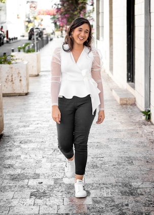 A woman in a soft pastel blouse and white pants, smiling while walking through a sunlit street