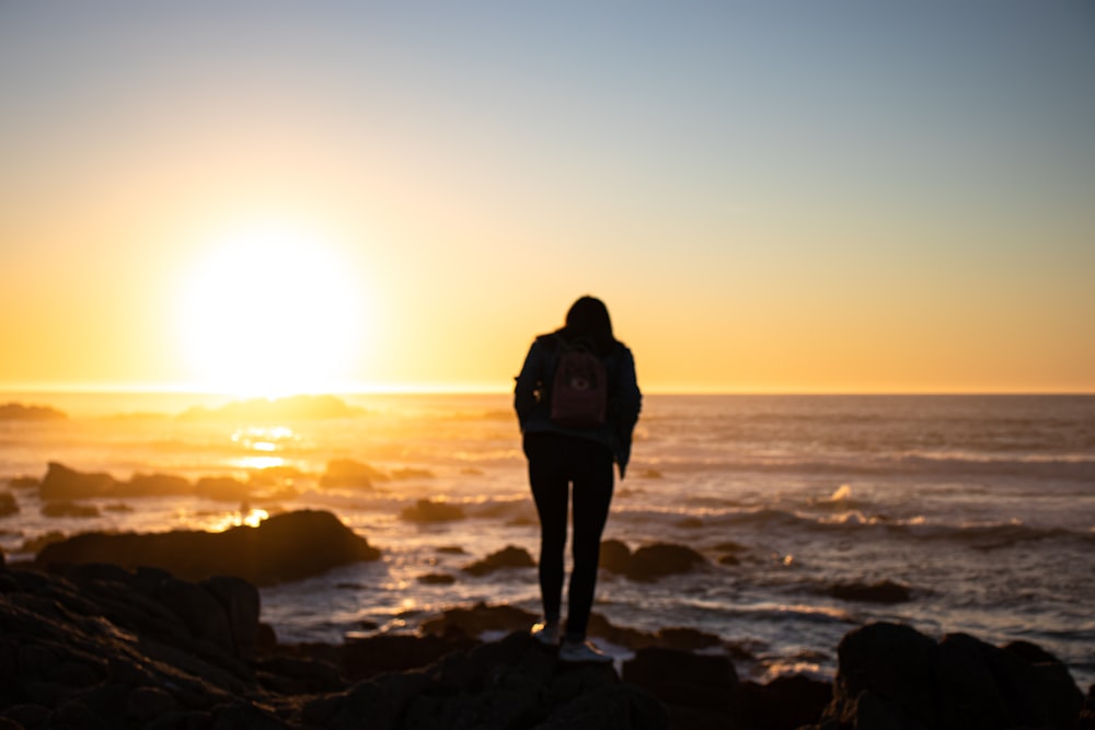 homem na jaqueta preta de pé na formação rochosa perto do mar durante o pôr do sol