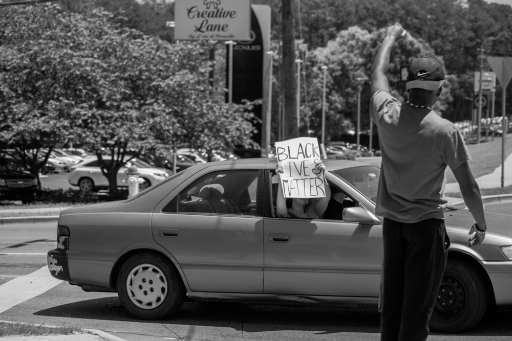 grayscale photo of man in black shirt and pants standing beside car