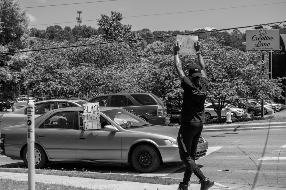 grayscale photo of man in black t-shirt and pants standing beside car