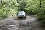 A Mitsubishi Triton splashing through a muddy forest trail with lush greenery around.