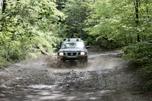 A Mitsubishi Triton splashing through a muddy forest trail with lush greenery around.