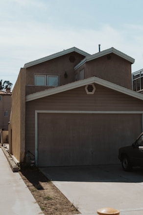 A two-story residential house with a flat, brown exterior. The building features a large double garage door and two small windows with shutters above. The driveway is a partially paved path leading to the street, with a dark vehicle partially visible on the right. The sky in the background is clear and blue.