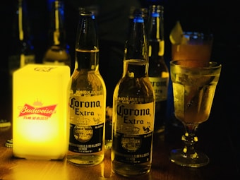 Several beer bottles of Corona Extra are placed on a wooden surface. A glowing Budweiser sign adds a warm, yellow light to the scene, along with a cocktail glass filled with a drink garnished with a lime. The ambiance suggests a dimly lit bar or lounge setting.