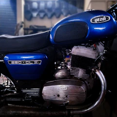 A vintage motorcycle with a blue fuel tank and side cover, featuring the brand name and model info. The engine and exhaust system are prominently visible, showing metallic components with a polished finish. The motorcycle appears to be stationary in a dimly lit garage or workshop with blurred background elements.