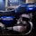 A vintage motorcycle with a blue fuel tank and side cover, featuring the brand name and model info. The engine and exhaust system are prominently visible, showing metallic components with a polished finish. The motorcycle appears to be stationary in a dimly lit garage or workshop with blurred background elements.