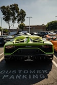 A bright green sports car, specifically a Lamborghini, is parked in a designated customer parking space. The car's bold design features a noticeable rear spoiler and distinct exhaust pipes. The parking lot is filled with other vehicles, and trees and a building are visible in the background under a clear sky.