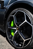 A close-up of a luxury sports car's wheel, featuring a sleek black rim with a distinctive geometric design and a bright green brake caliper. The tire is a Pirelli P-Zero, highlighting performance and style.