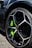A close-up of a luxury sports car's wheel, featuring a sleek black rim with a distinctive geometric design and a bright green brake caliper. The tire is a Pirelli P-Zero, highlighting performance and style.