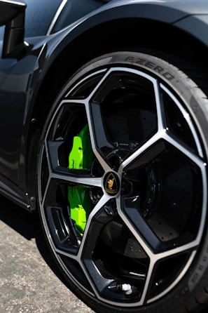 A close-up of a luxury sports car's wheel, featuring a sleek black rim with a distinctive geometric design and a bright green brake caliper. The tire is a Pirelli P-Zero, highlighting performance and style.
