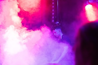 A person stands amidst colorful mist and vibrant stage lighting. The atmosphere is dynamic, with hues of pink, purple, and white creating an otherworldly scene. Lighting equipment is partially visible, suggesting a live performance or event setting.