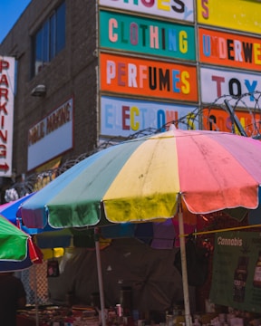A street market scene features a colorful umbrella with segments in yellow, green, blue, and red. Behind it, a large building facade is visible with bold, brightly colored signage advertising various products like clothing, perfumes, and toys. The environment appears urban, with visible barbed wire atop a fence, and a sign mentioning cannabis products.