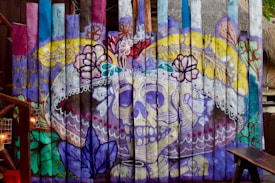 A colorful mural painted on vertical wooden planks depicts a stylized skull adorned with floral patterns and intricate designs in vibrant hues. The artwork incorporates vivid flowers and bold outlines, giving it a festive and lively appearance.