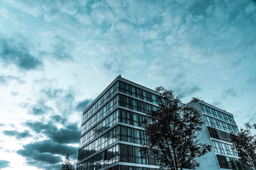 A modern glass-fronted building with multiple floors is shown against a backdrop of an expansive sky filled with clouds. Trees are visible in the foreground, partially obscuring the lower parts of the building.