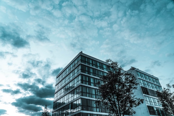 A modern glass-fronted building with multiple floors is shown against a backdrop of an expansive sky filled with clouds. Trees are visible in the foreground, partially obscuring the lower parts of the building.