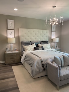 A modern bedroom features a large upholstered headboard with tufted design behind a neatly made bed with multiple pillows. Two bedside tables, each with a lamp and decorative items, flank the bed. A contemporary chandelier hangs from the ceiling, providing ambient light. The decor includes framed artwork on the walls above the lamps. A small cushioned bench sits at the foot of the bed, completing the tranquil and sophisticated design.