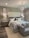 A modern bedroom features a large upholstered headboard with tufted design behind a neatly made bed with multiple pillows. Two bedside tables, each with a lamp and decorative items, flank the bed. A contemporary chandelier hangs from the ceiling, providing ambient light. The decor includes framed artwork on the walls above the lamps. A small cushioned bench sits at the foot of the bed, completing the tranquil and sophisticated design.