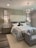A modern bedroom features a large upholstered headboard with tufted design behind a neatly made bed with multiple pillows. Two bedside tables, each with a lamp and decorative items, flank the bed. A contemporary chandelier hangs from the ceiling, providing ambient light. The decor includes framed artwork on the walls above the lamps. A small cushioned bench sits at the foot of the bed, completing the tranquil and sophisticated design.