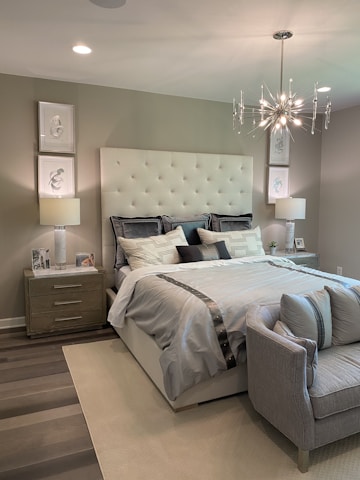 A modern bedroom features a large upholstered headboard with tufted design behind a neatly made bed with multiple pillows. Two bedside tables, each with a lamp and decorative items, flank the bed. A contemporary chandelier hangs from the ceiling, providing ambient light. The decor includes framed artwork on the walls above the lamps. A small cushioned bench sits at the foot of the bed, completing the tranquil and sophisticated design.