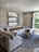 A modern living room featuring a large, plush sectional sofa in neutral tones with several decorative pillows in geometric and solid patterns. Above the sofa, three horizontal rectangular mirrors with metallic frames are mounted on a textured wall. A distinctive wooden coffee table with a natural, organic design sits on a round gray rug. A sleek chandelier with candle-style lights is suspended from the ceiling. Large windows with striped curtains allow natural light to fill the room, offering a view of greenery outside.