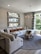 A modern living room featuring a large, plush sectional sofa in neutral tones with several decorative pillows in geometric and solid patterns. Above the sofa, three horizontal rectangular mirrors with metallic frames are mounted on a textured wall. A distinctive wooden coffee table with a natural, organic design sits on a round gray rug. A sleek chandelier with candle-style lights is suspended from the ceiling. Large windows with striped curtains allow natural light to fill the room, offering a view of greenery outside.