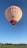 A large hot air balloon with 'Getzner Textil AG' and a logo printed on its surface is floating in the clear sky. The balloon has a white color with red and dark blue details. A wicker passenger basket is attached to the balloon, and a scenic landscape of green fields and distant hills is visible below.