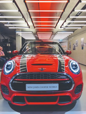 A bright red sports car is displayed under modern lighting, showcasing its sleek design and distinctive front grille. The lighting creates a dramatic effect, enhancing the car's glossy finish.