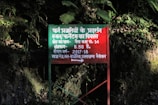 A red rectangular signboard with text in Hindi is situated against a background of dense foliage. The area is lush with green leaves and shadows suggest it's in a forested or heavily vegetated area.