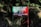A red rectangular signboard with text in Hindi is situated against a background of dense foliage. The area is lush with green leaves and shadows suggest it's in a forested or heavily vegetated area.