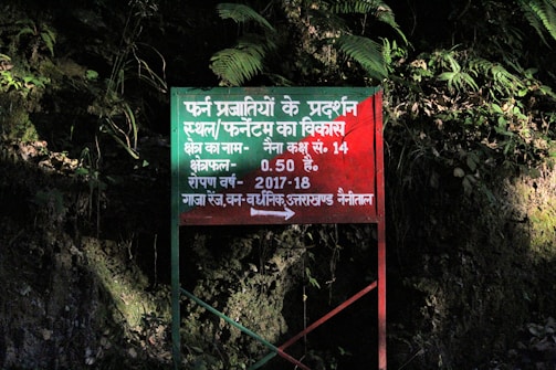A red rectangular signboard with text in Hindi is situated against a background of dense foliage. The area is lush with green leaves and shadows suggest it's in a forested or heavily vegetated area.