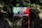 A red rectangular signboard with text in Hindi is situated against a background of dense foliage. The area is lush with green leaves and shadows suggest it's in a forested or heavily vegetated area.