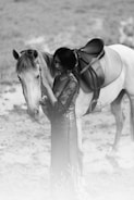 A woman stands beside a white horse in a natural setting. She is wearing a long, elegant dress with intricate patterns. Her right hand rests gently on the horse's face, showcasing a close bond between the two. The background is a soft blur, focusing the attention on the woman and the horse.