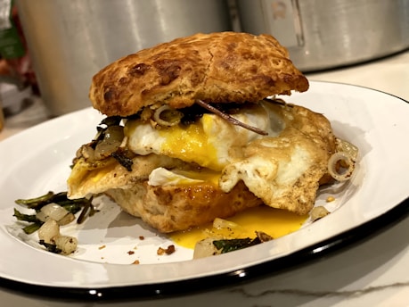 A close-up view of a sandwich featuring a flaky biscuit, fried eggs with runny yolks, and sautéed onions and greens. The sandwich is placed on a white plate with a black rim, on what appears to be a kitchen countertop background with metal containers partially visible.