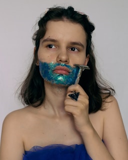 A woman with shoulder-length hair is holding a razor to her face. Her cheeks and chin are covered with bright blue and green glitter, suggesting a creative, artistic expression. She is wearing a strapless blue dress and a large triangular ring. The background is neutral, putting focus on her and the vivid colors on her face.